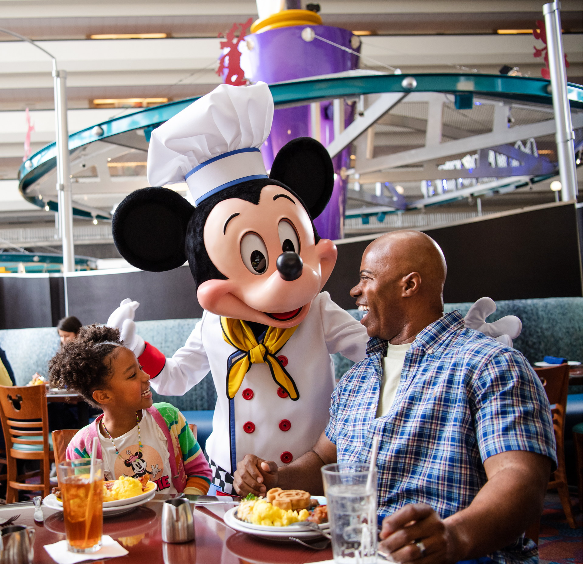 Mickey en toque de chef salue une fillette et son père attablés devant leur assiette, sous le rail du monorail, au Chef Mickey’s