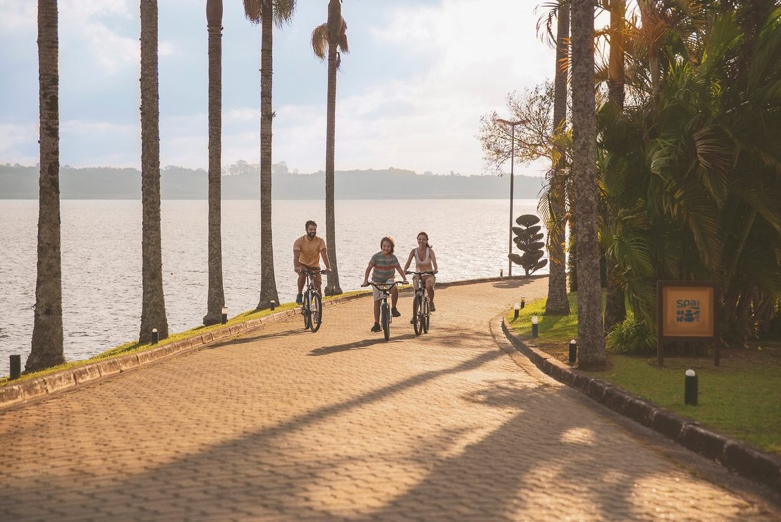 Family cycling at Club Med Lake Paradise, Brazil, along the lake, surrounded by palm trees and lush greenery.