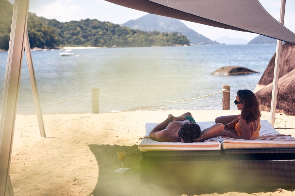 Couple lounging on a shaded beach bed at Club Med Rio Das Pedras, enjoying a serene ocean view.