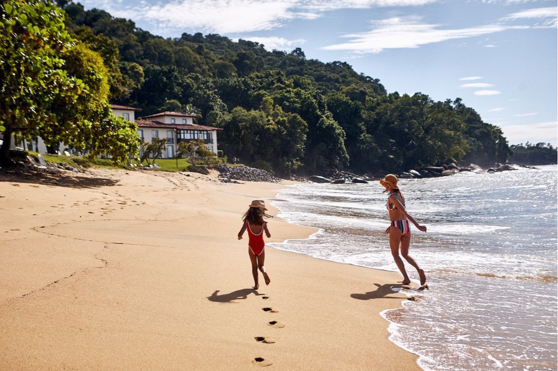 Femme et enfant marchant sur la plage au Club Med Rio Das Pedras, avec le complexe et une forêt luxuriante en arrière-plan.