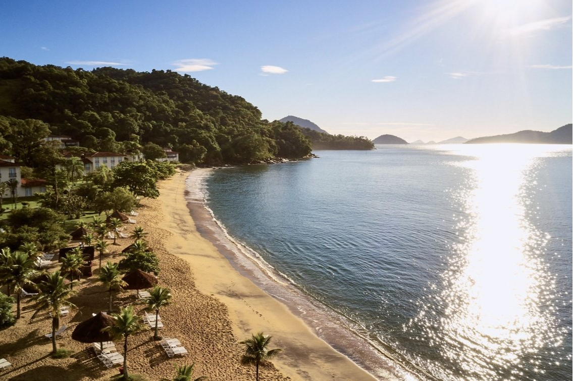 Aerial view of Club Med Rio Das Pedras with its palm-lined beach, lounge chairs, and a mountain backdrop.