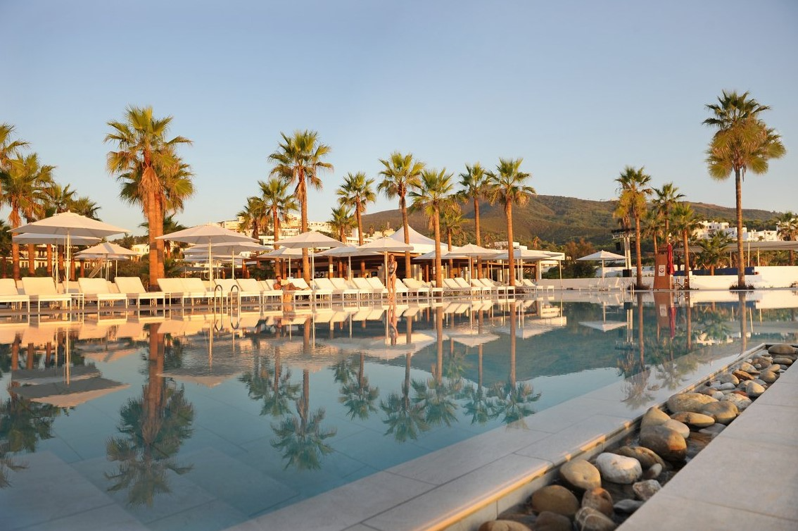 Vue de la piscine au Club Med Yasmina, entourée de chaises longues, parasols, et palmiers, pour un moment de relaxation.