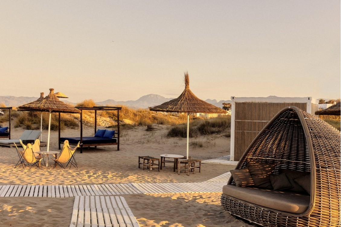 Vue de la plage au Club Med Yasmina, avec lits de détente, montagnes en arrière-plan et chemin en bois dans le sable.