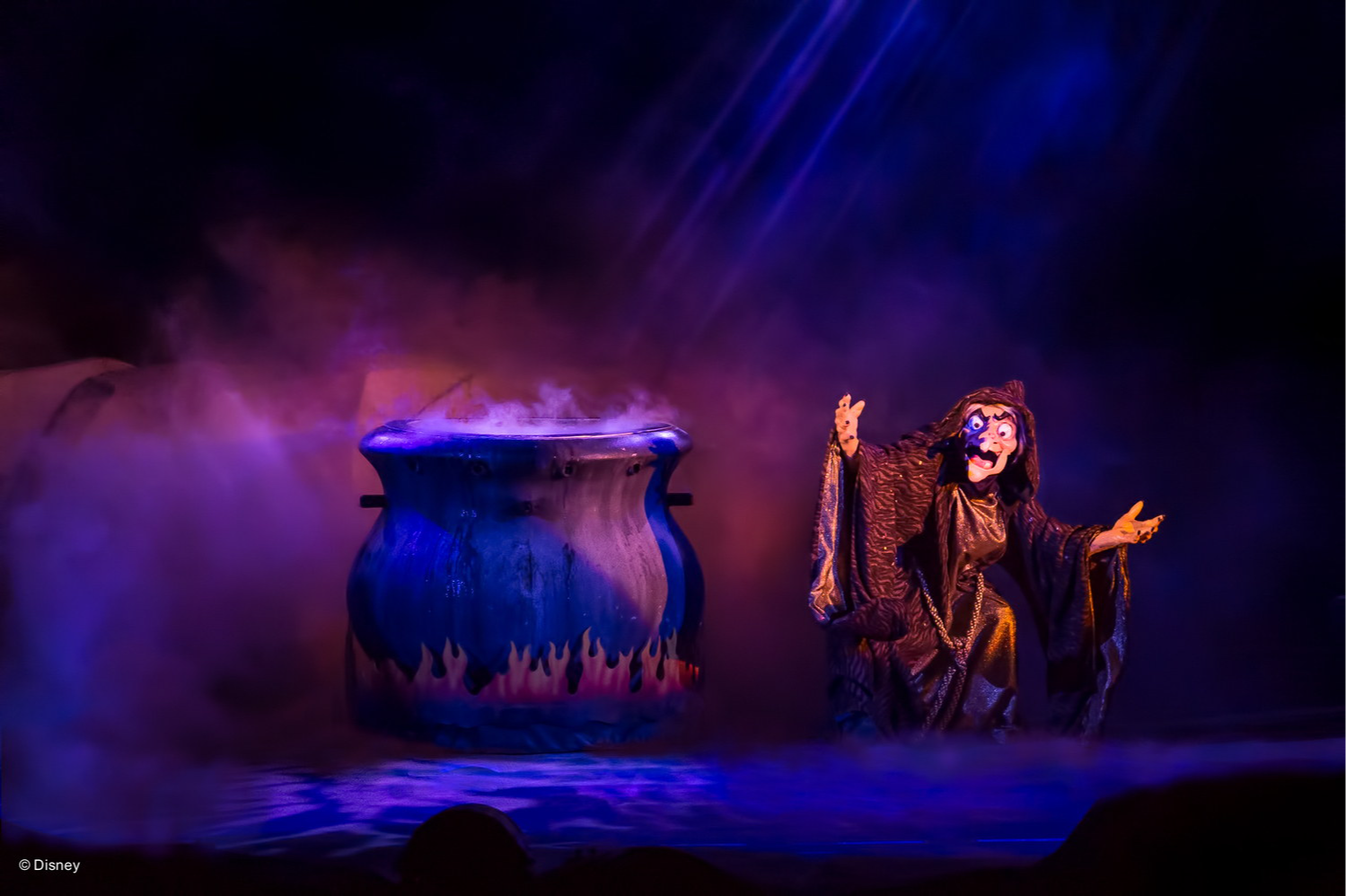 The witch from Snow White, arms outstretched beside a giant cauldron in purple smoke, during the Fantasmic show, at Disney’s Hollywood Studios
