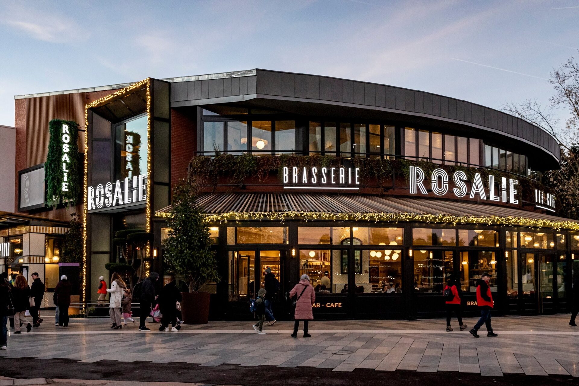 Façade illuminée de la Brasserie Rosalie à Disney Village au crépuscule, avec des passants devant sa verrière. Photo : © Rosalie / Simon Detraz.