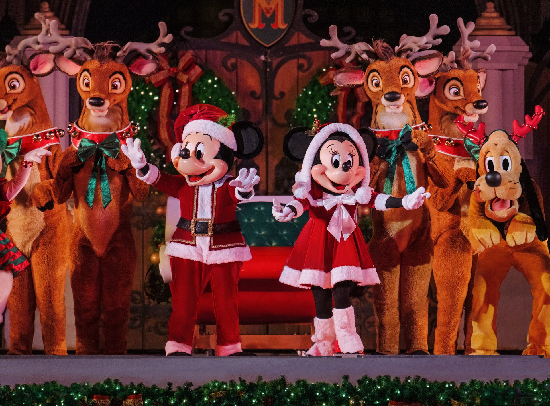 Mickey et Minnie en habits de père Noël saluent depuis un traîneau entouré de rennes costumés, sur une scène décorée de guirlandes, à Magic Kingdom