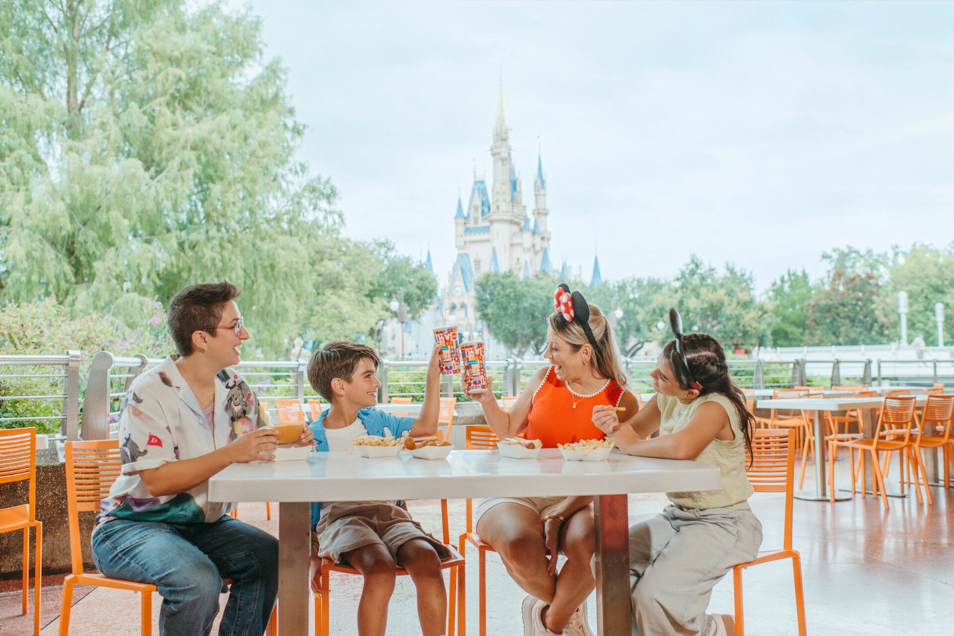 Quatre convives trinquent autour d’une table de terrasse orange, le château de Cendrillon au loin derrière les arbres, à Magic Kingdom