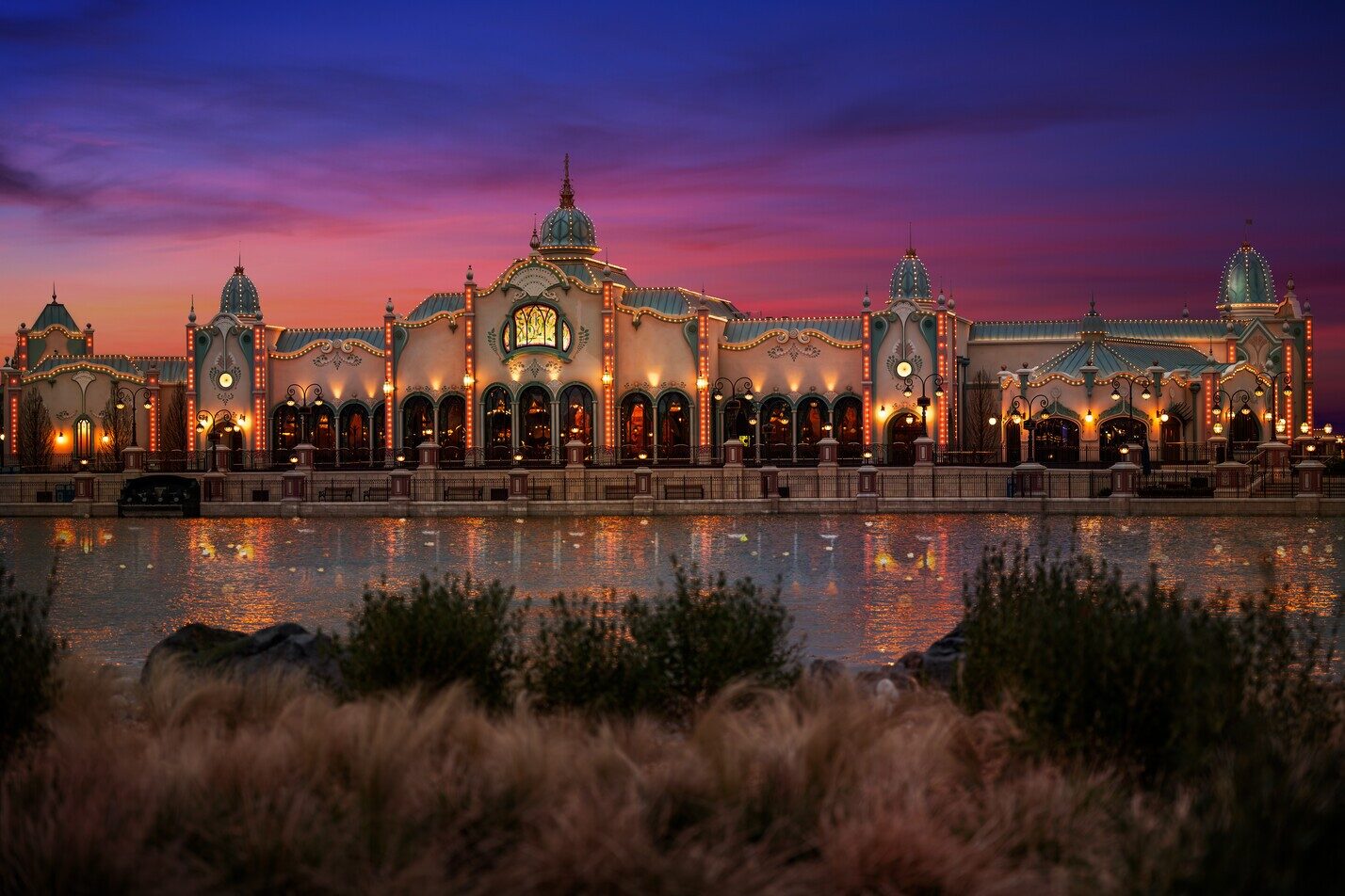 The lit pavilions of Disney Adventure World at dusk, reflected in Adventure Bay beneath a pink and purple sky. Photo: ©Disney.