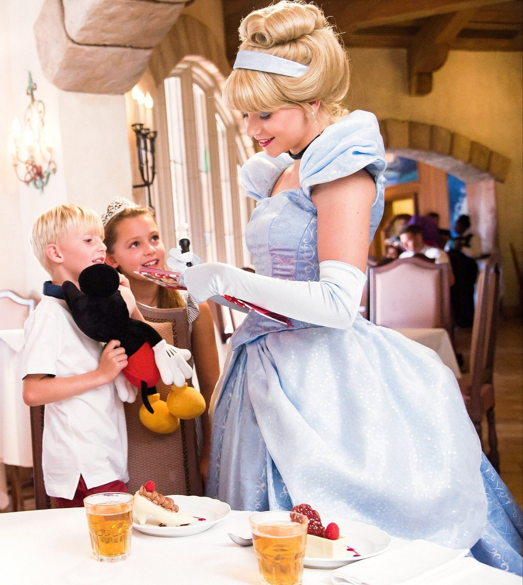 Cendrillon salue deux enfants attablés à l'Auberge de Cendrillon, au Parc Disneyland, dont l'un serre une peluche de Mickey. Photo : ©Disney. Cendrillon salue deux enfants attablés à l'Auberge de Cendrillon, au Parc Disneyland, dont l'un serre une peluche de Mickey. Photo : ©Disney.
