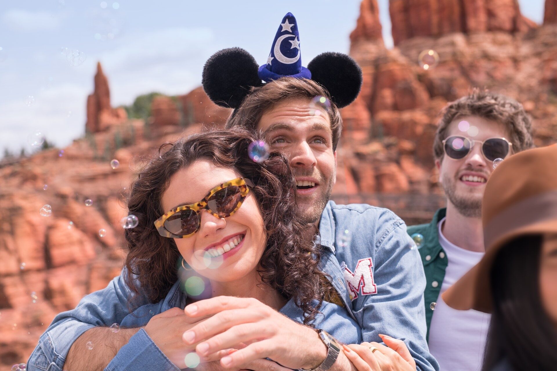 Two smiling guests, one wearing a Mickey ears hat, in front of the red rocks of Big Thunder Mountain. Photo: ©Disney. Two smiling guests, one wearing a Mickey ears hat, in front of the red rocks of Big Thunder Mountain. Photo: ©Disney.