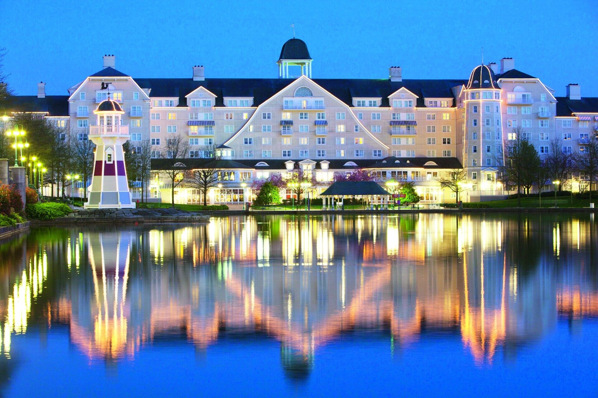 Le Disney Newport Bay Club illuminé au crépuscule, avec son phare rayé et sa longue façade qui se reflètent dans le lac Disney. Photo : ©Disney.