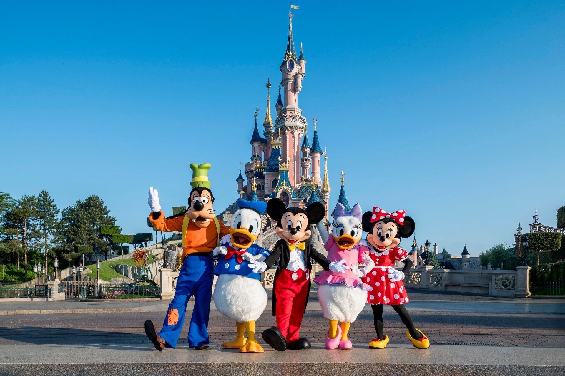 Dingo, Donald, Mickey, Daisy et Minnie devant le Château de la Belle au Bois Dormant, au Parc Disneyland, sous un ciel bleu. Photo : ©Disney. Dingo, Donald, Mickey, Daisy et Minnie devant le Château de la Belle au Bois Dormant, au Parc Disneyland, sous un ciel bleu. Photo : ©Disney.