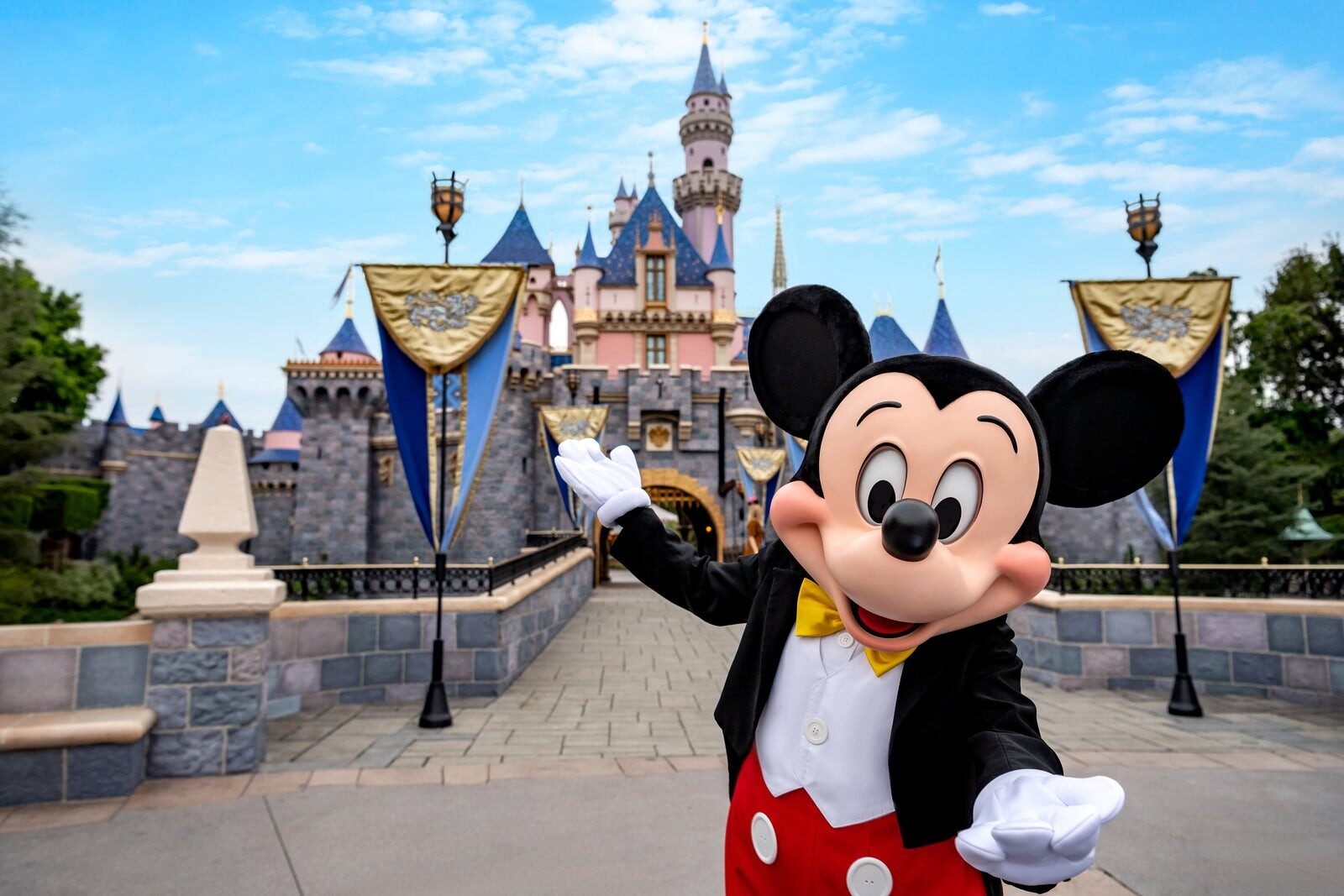 Mickey Mouse waving in front of Sleeping Beauty Castle and its blue banners