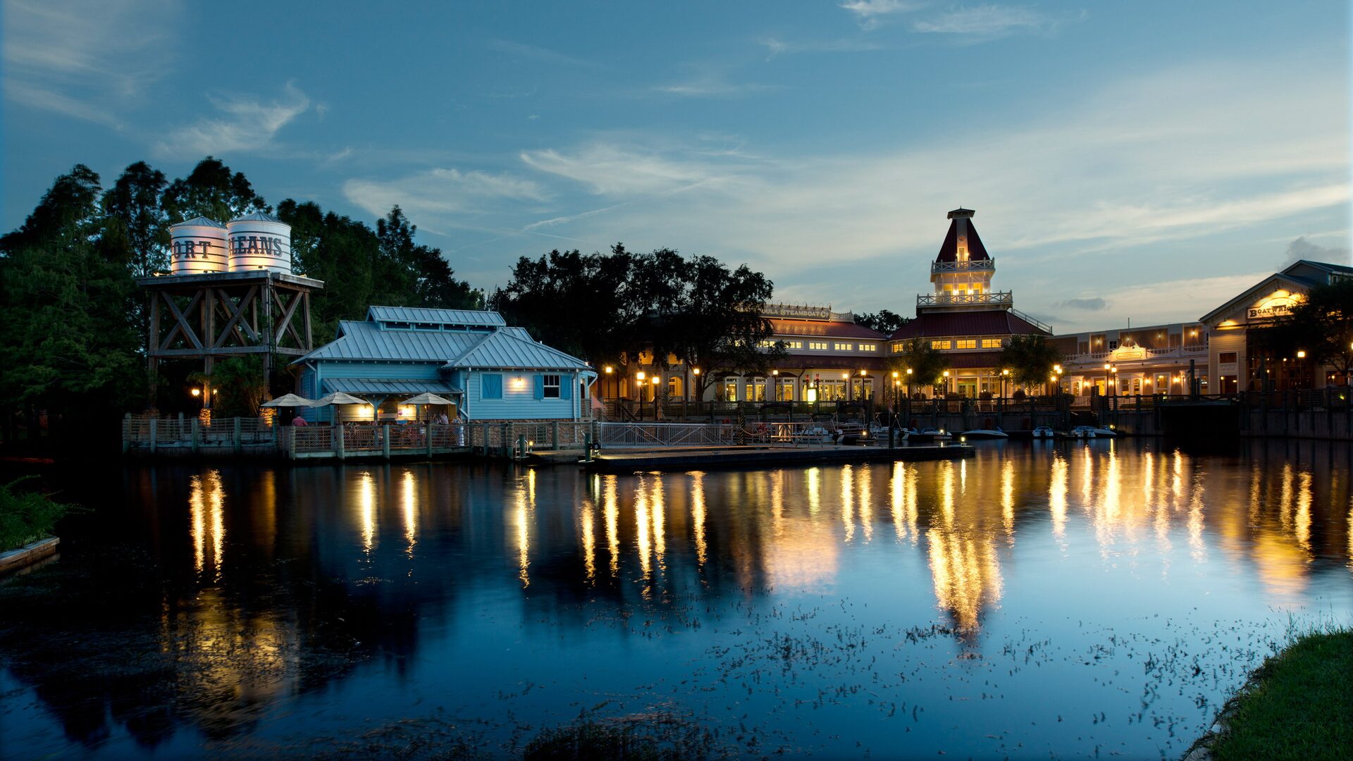 The lit buildings of Disney’s Port Orleans Resort – Riverside reflected in the river at dusk, the water tower on the left