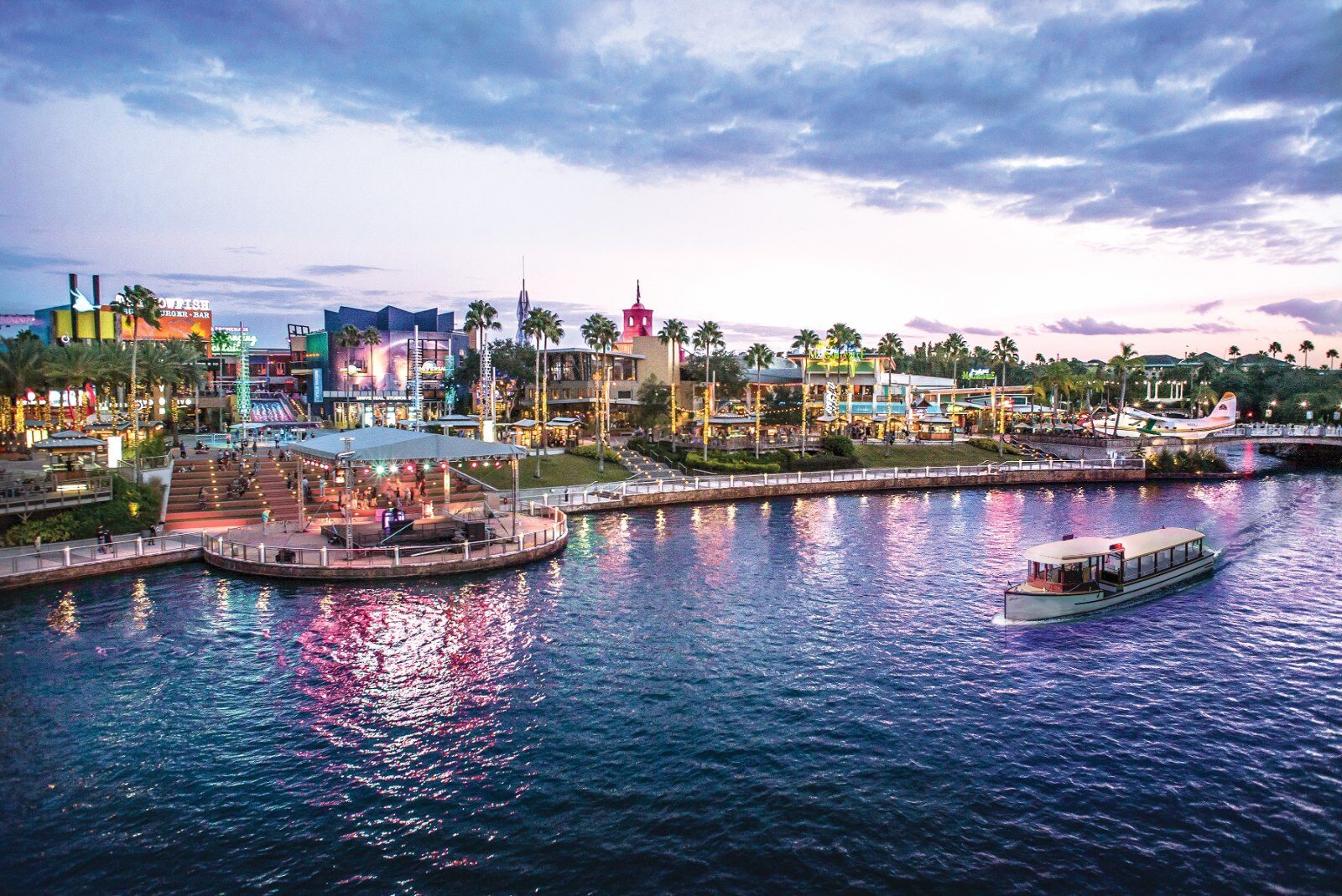Universal CityWalk at dusk seen across the lagoon, with the amphitheatre, restaurant neon and a water taxi